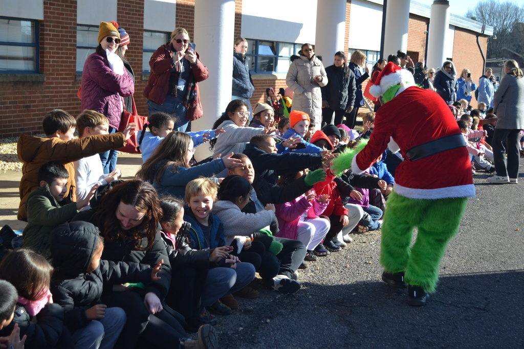 Image contains students and teachers joyfully watching  a Grinch character give high fives as he walks by students.  Everyone appears happy and some are filming with their phones.