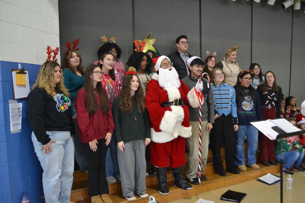 Image contains the high school choir singing Christmas songs in festive dress in the elementary school gym.  Singers have on headbands.  Boy in center is dressed as Santa.