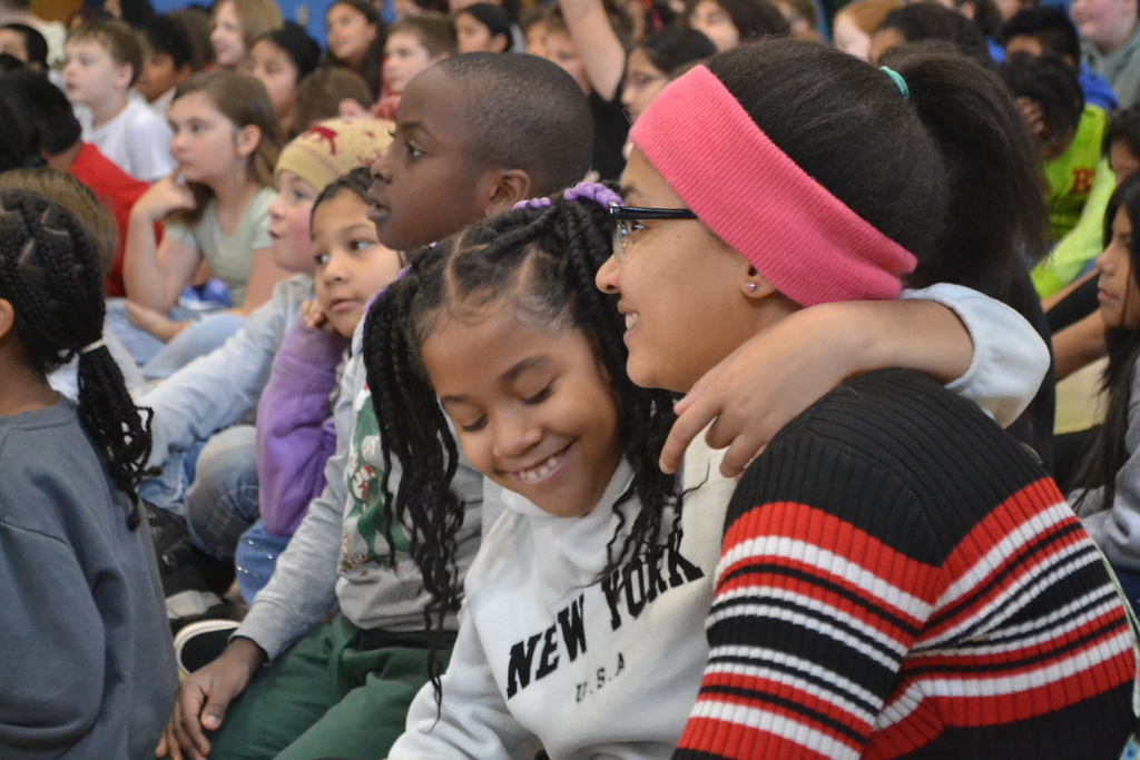 Image contains a staff member smiling while a student in a NewYork Sweatshirt hugs and smiles during the concert in the gym.