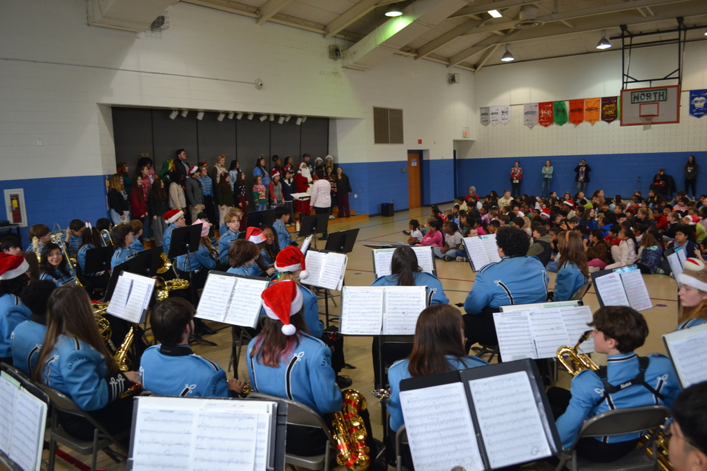 Semi arial image taken from behind the music stands of the band while the choir performs on side stairs while elementary students listen.