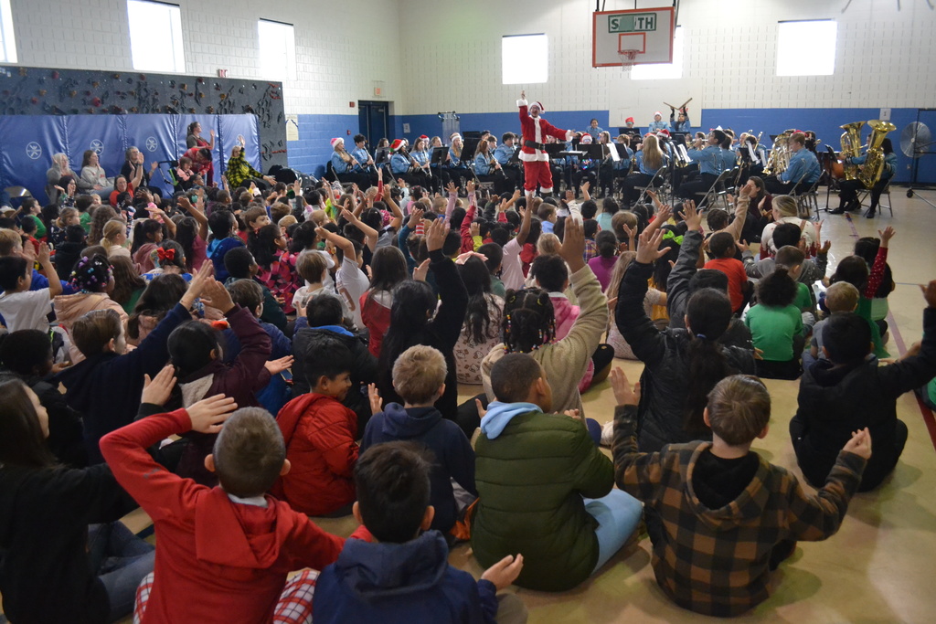 Image contains members of the band in blue uniforms playing their instruments at a winter themed concert in the elementary school gym.  Students are clapping and cheering along.