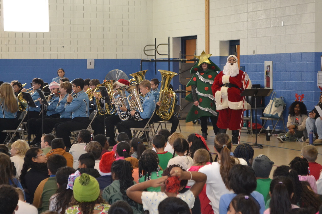 Image contains members of the band in blue uniforms playing their instruments at a winter themed concert in the elementary school gym.