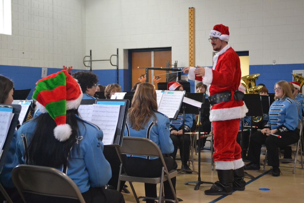 Image contains members of the band in blue uniforms playing their instruments at a winter themed concert in the elementary school gym.