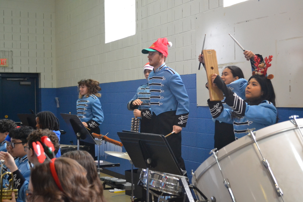 Image contains members of the band in blue uniforms playing their instruments at a winter themed concert in the elementary school gym.