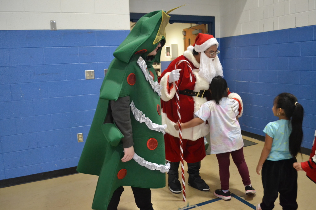 Image contains a high school student dressed as Santa and a Christmas Tree saying goodbye as a line of elementary students leaves the gym.