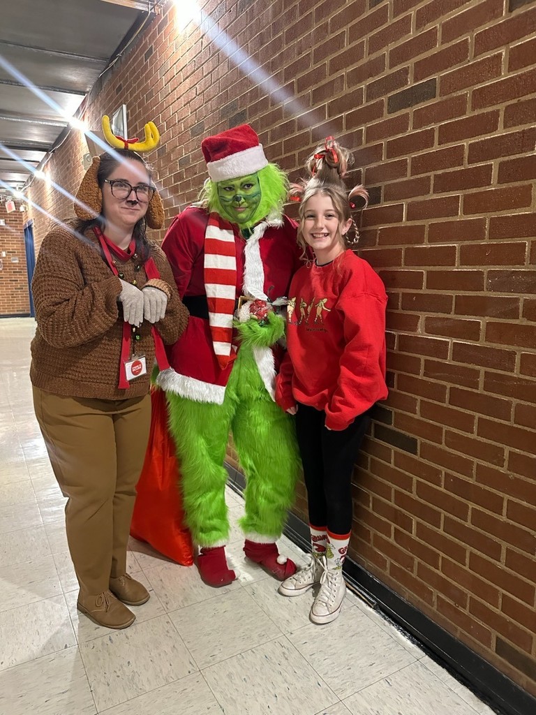Three people pose in a school hallway for Grinch Spirit Day at Culpeper Middle School. One person is dressed as the Grinch in a Santa-style outfit with green fur, one is dressed as Cindy Lou Who in a red holiday sweatshirt with festive hair accessories, and one is dressed as Max the dog wearing reindeer antlers. All are smiling and standing against a brick wall.