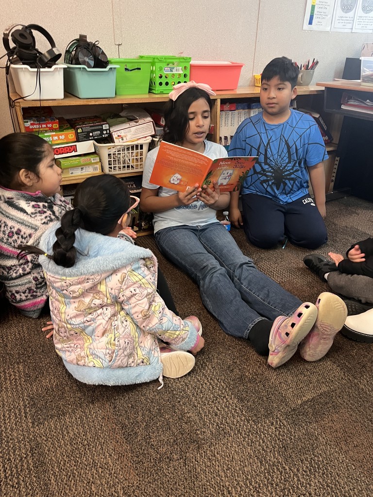 Two 4th grade students read to two preschool students in the classroom on the floor.