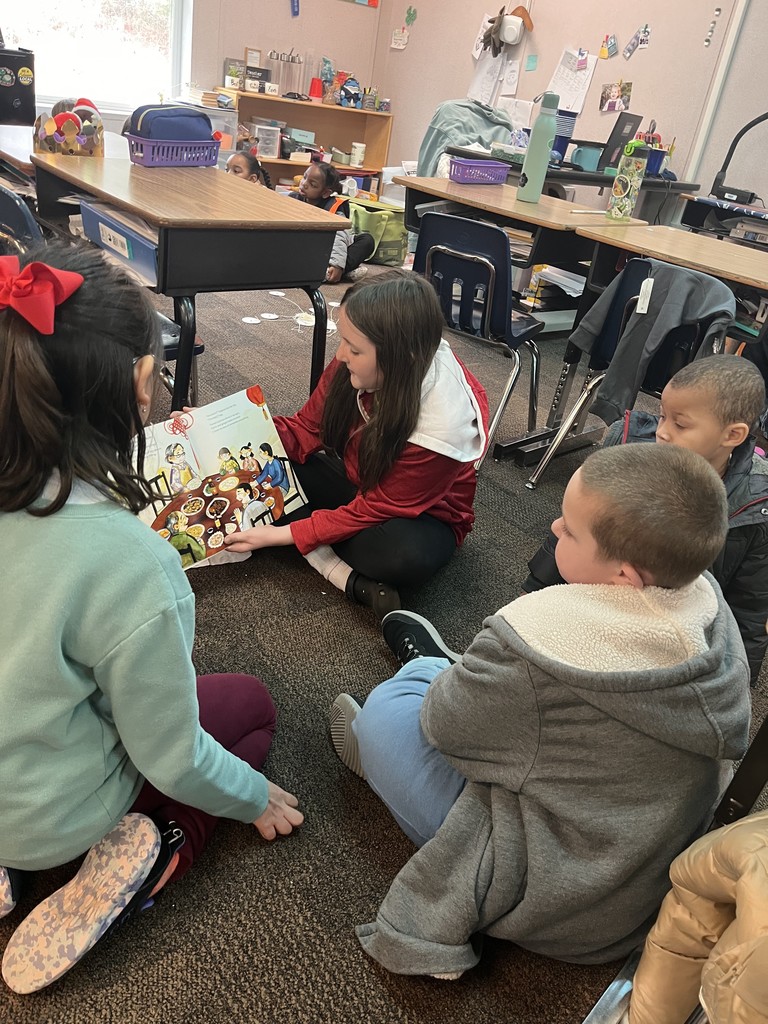Two 4th grade girls read to two preschool students in the classroom on the floor.