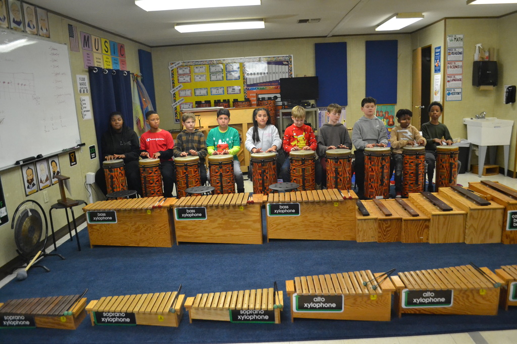 10 students sit behind ORFF drums in a music classroom ready to perform.