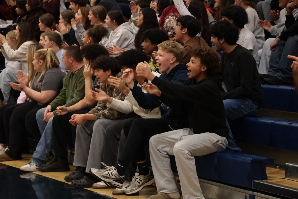 Students sitting in the front row of bleachers clap animatedly.
