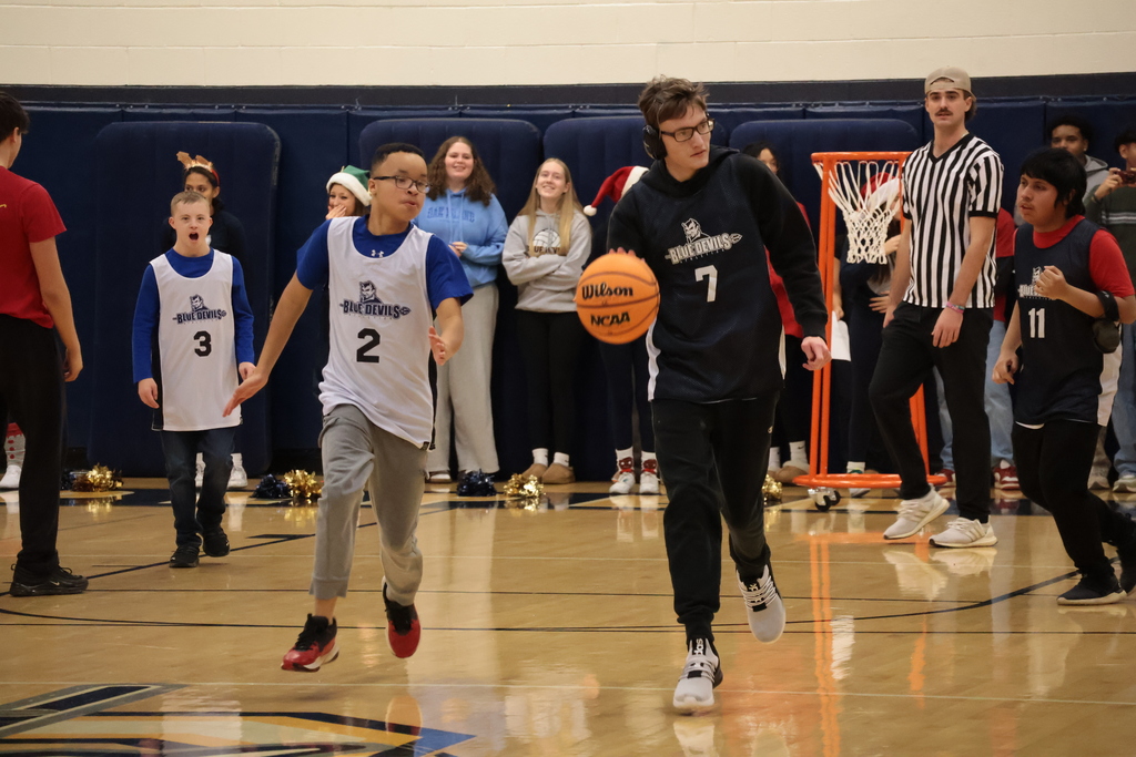 A basketball player in a navy jersey dribbles down the court. He is followed by an opposing player in a white jersey.