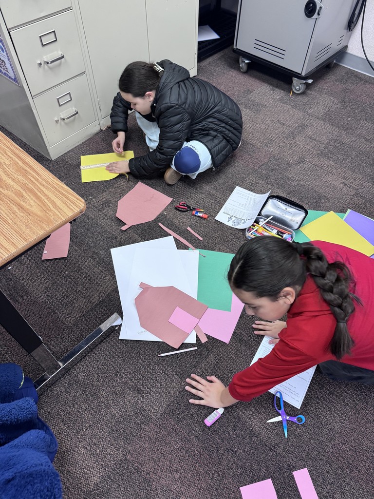 Two girls are working on their classroom floor with a variety of colored paper to make their Gingerbread Math Houses.