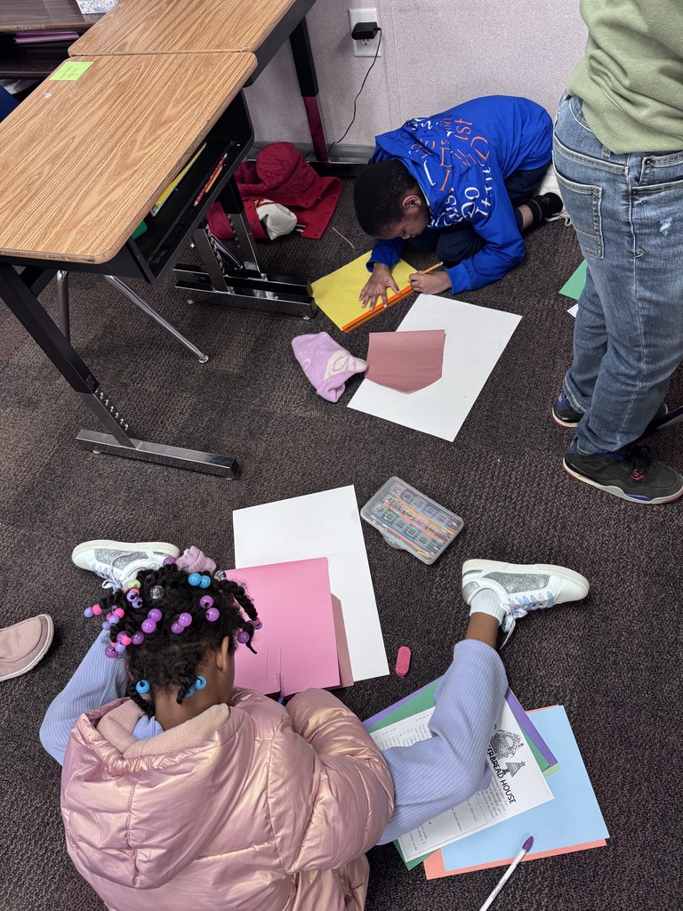 Two students are working on their classroom floor with a variety of colored paper to make their Gingerbread Math Houses.