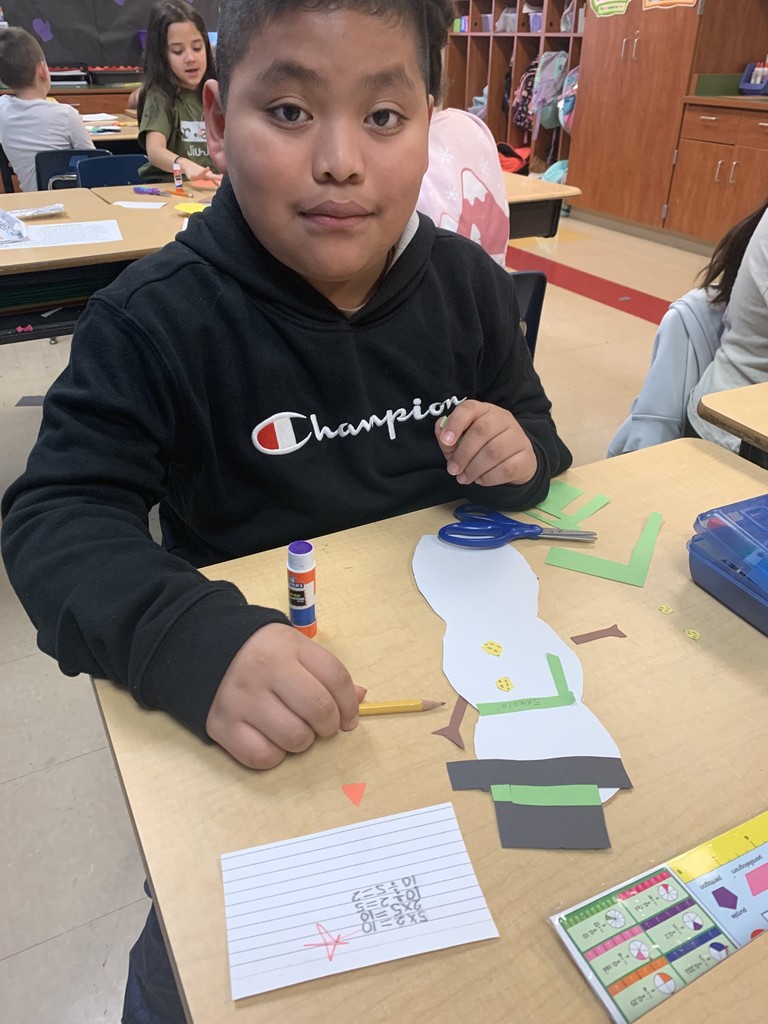 A student sits at a classroom table working on a paper snowman craft. The student holds a pencil near the snowman cutout, which has paper decorations and math facts written on it. Scissors, glue, and paper pieces are spread across the desk.