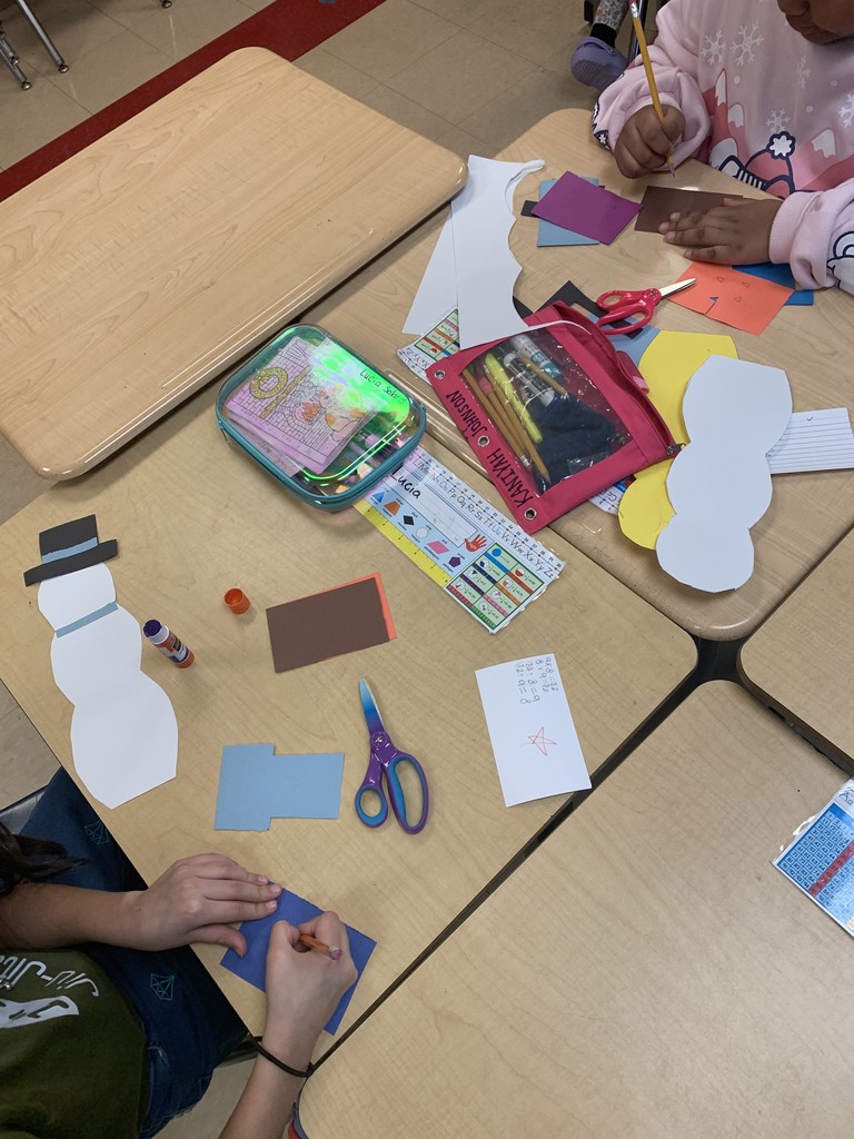 An overhead view of classroom desks covered with snowman craft materials, including white paper snowman cutouts, colored construction paper, scissors, glue sticks, and pencil cases.