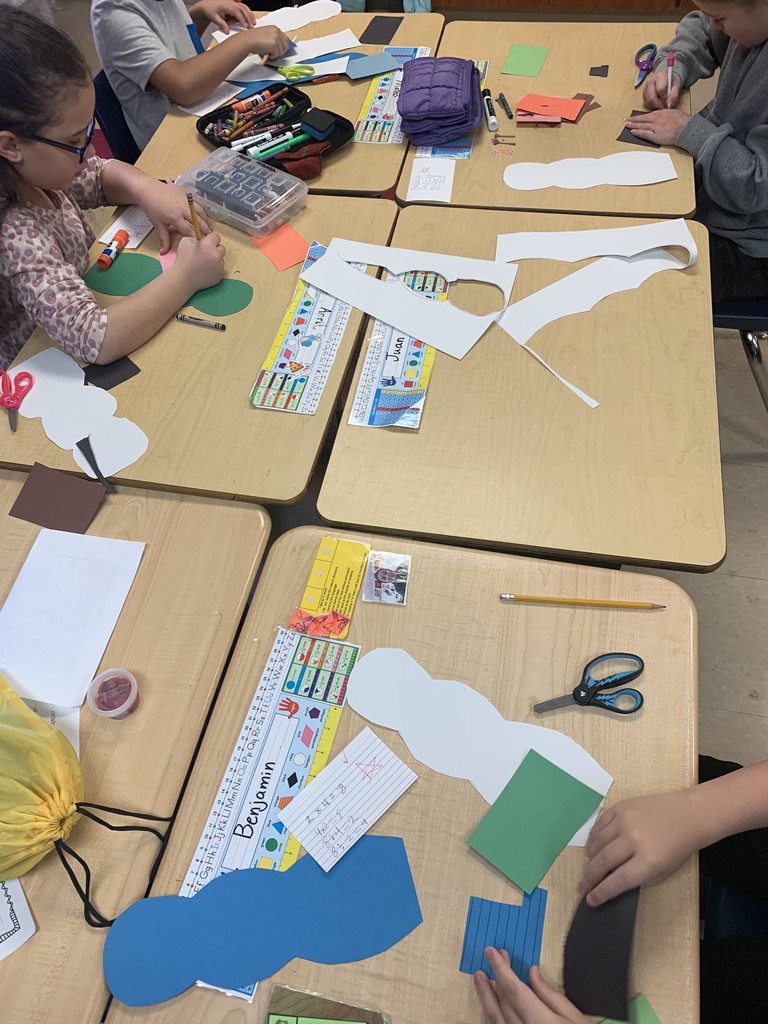Multiple students work at grouped desks, cutting and decorating paper snowman shapes. Math cards, rulers, and art supplies are spread across the tables as students focus on their projects.
