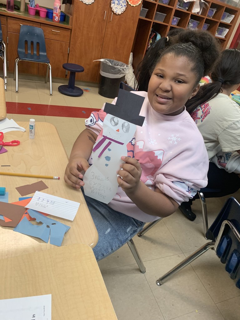 A smiling student holds up a completed paper snowman decorated with a black hat, scarf, and buttons. Math equations are written on the snowman’s body. Art supplies and paper scraps are visible on the desk.