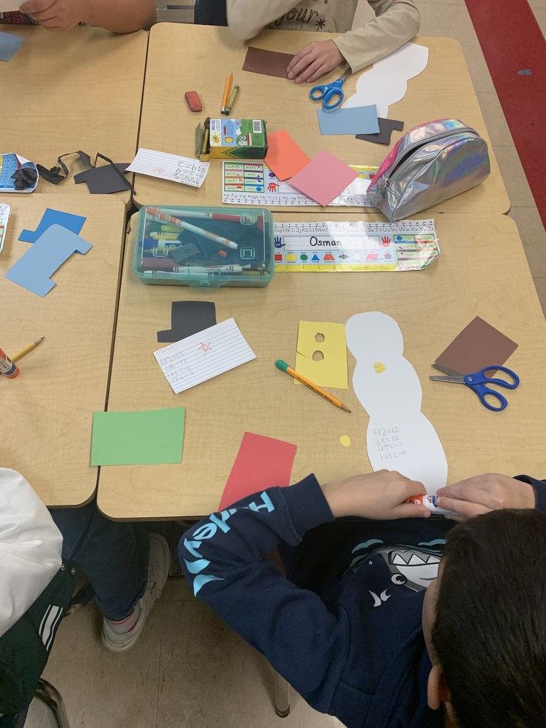 A close overhead view of desks showing partially completed snowman crafts with math facts written on them. Scissors, glue sticks, colored paper, and name labels are visible.