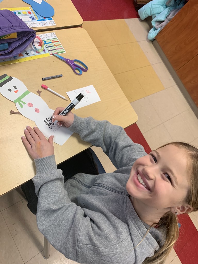 A student smiles up at the camera while writing math facts on a paper snowman using a marker. The snowman craft lies flat on the desk, surrounded by scissors, crayons, and paper pieces.