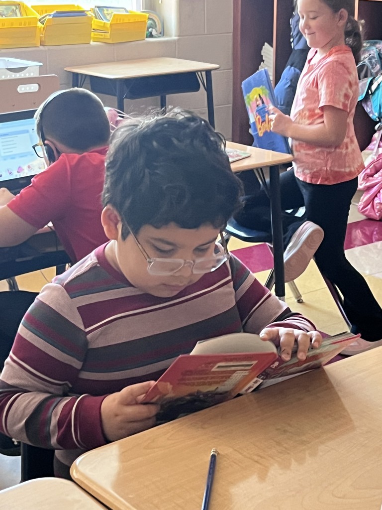 A student wearing glasses reads a book at their desk during choice time while other classmates read and work independently around the classroom.