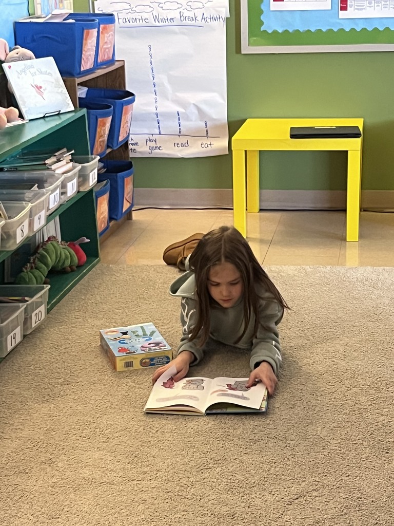 A student lies comfortably on the classroom carpet reading a picture book during choice time, with classroom shelves, bins, and a student-made graph visible in the background.