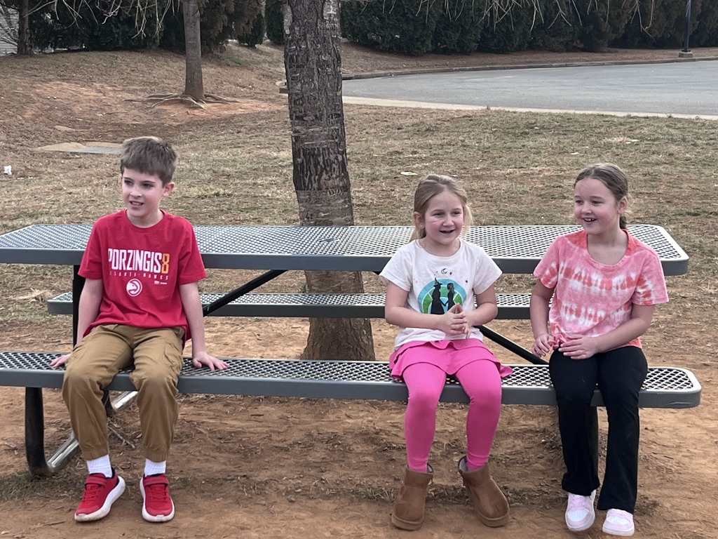 Three students sit together on a picnic bench outside during recess, smiling and talking while enjoying the mild weather.