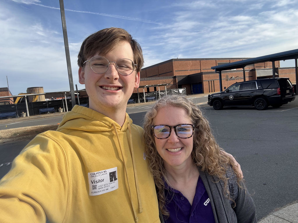 A smiling young man wearing glasses and a yellow hoodie with a Culpeper Middle School visitor badge takes a selfie with a smiling adult woman wearing glasses and a purple CMS shirt. They are standing outside a school building on a sunny day, with the school and a parked sheriff’s vehicle visible in the background.