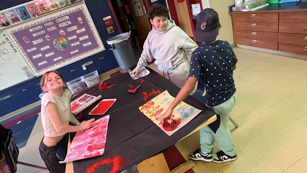 Four elementary students work together at an art table, rolling red and multicolored paint onto paper. One student smiles at the camera while others focus on their artwork in the brightly lit classroom.