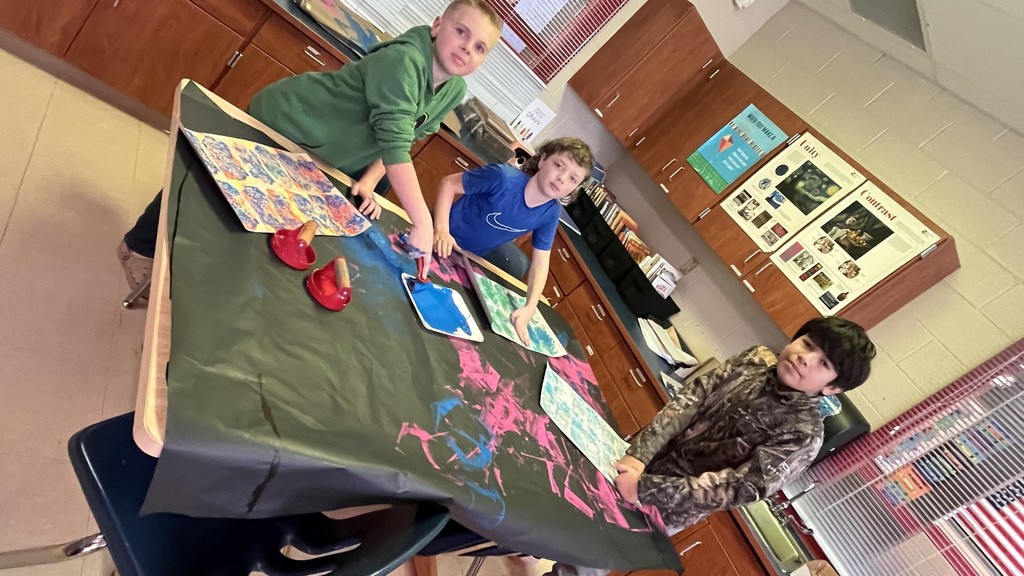 Three elementary students paint together at a table covered with black paper and red paint. One student presses paint onto paper while others prepare their prints, smiling and interacting during the art activity.