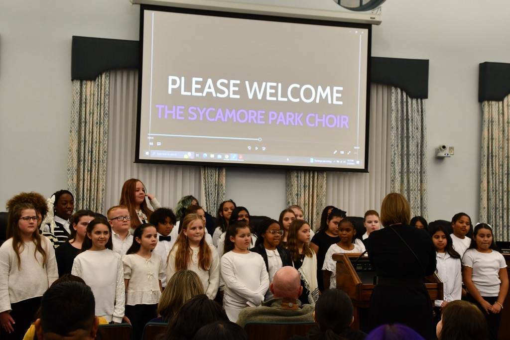 large group of students wearing white tops and black bottoms standing in the front of a board room under a screen that says Please Welcome the Sycamore Park Choir