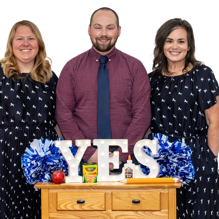 three adults smile for the camera while standing behind a small wooden table. on the table are the letters YES. also on the table are blue pom poms