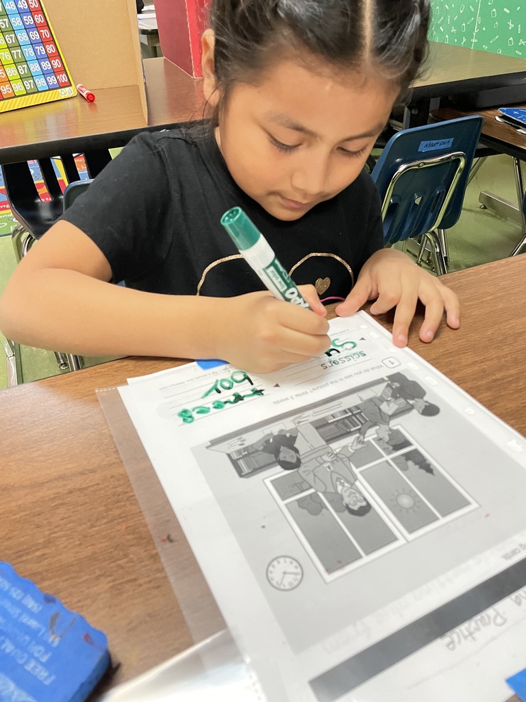A student concentrates while writing with a green marker on a worksheet that includes a classroom scene illustration.
