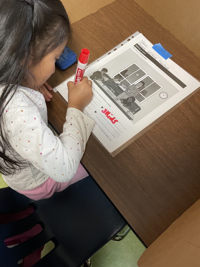 A young student uses a marker to label details on a picture-based writing worksheet while seated at a classroom desk.