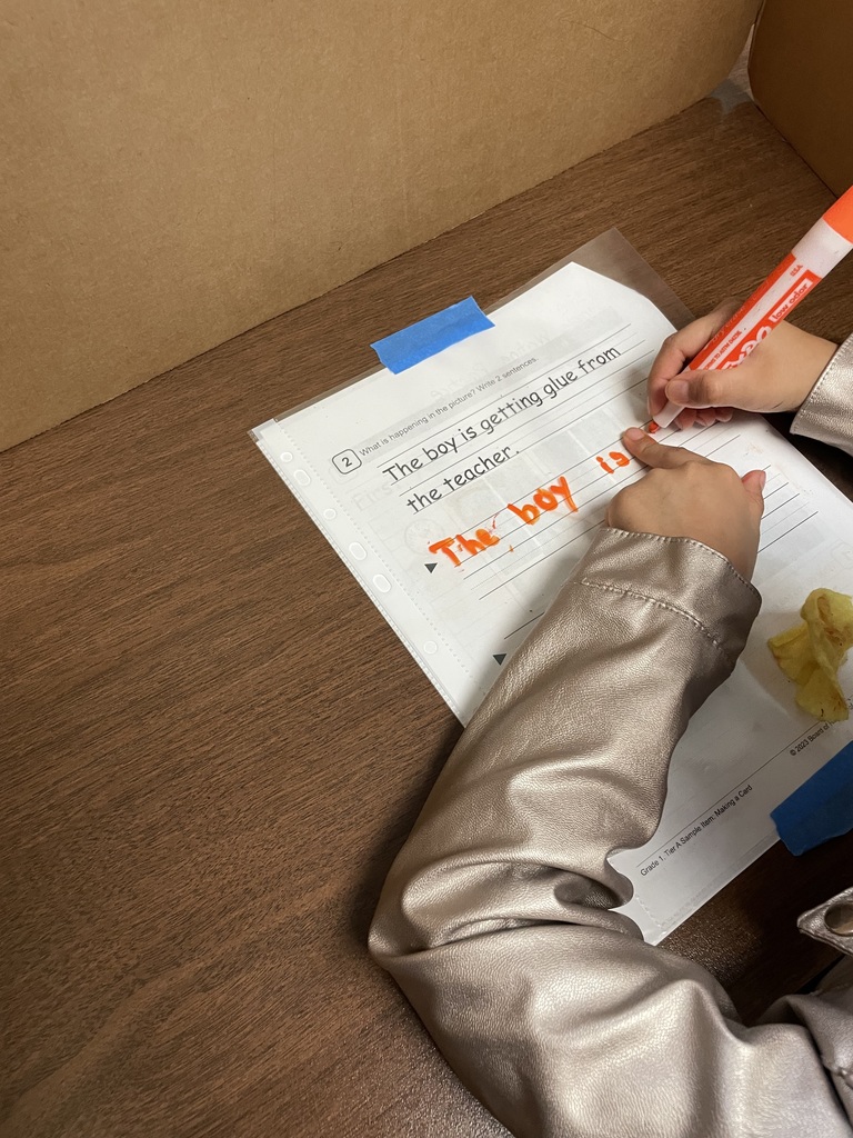 A student writes with a marker on a worksheet showing a picture prompt, practicing sentence writing at a desk with a privacy divider.