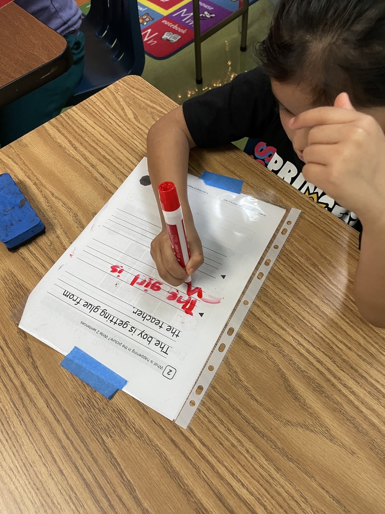 A student writes on a laminated worksheet with a marker, practicing identifying words based on a picture prompt.