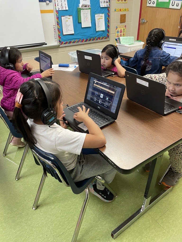 Several students wearing headphones work independently on laptops at a table, focusing on an online language activity in the classroom.