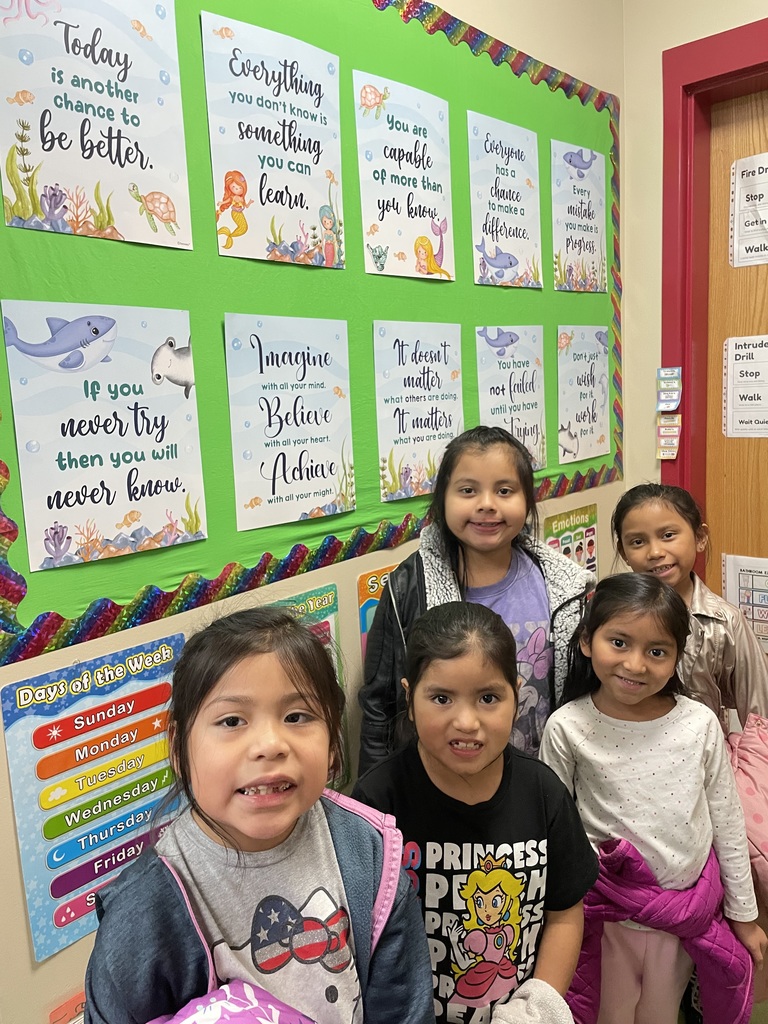 A small group of smiling students stands in front of a green bulletin board decorated with colorful motivational posters.