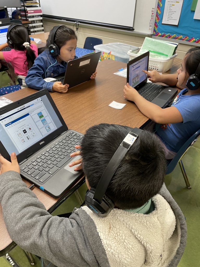 Students wearing headphones sit at a table using laptops, completing digital listening and language tasks together.
