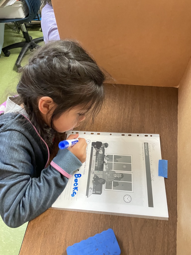 A student writes the word “books” on a picture-based worksheet while seated at a desk with a privacy divider.