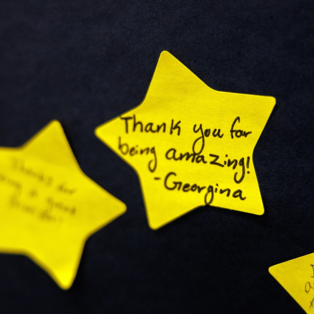 Close-up of a yellow, star-shaped note pinned to a dark bulletin board with a handwritten message that reads, ‘Thank you for being amazing!’ signed ‘Georgina.’ Other yellow star notes appear blurred in the background.