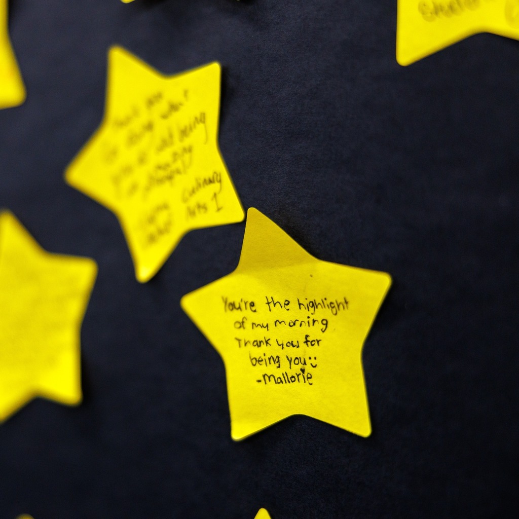 Close-up of a yellow, star-shaped note pinned to a bulletin board with a handwritten message that reads, ‘You’re the highlight of my morning. Thank you for being you. – Mallorie.’ Other yellow star notes are visible softly out of focus in the background.