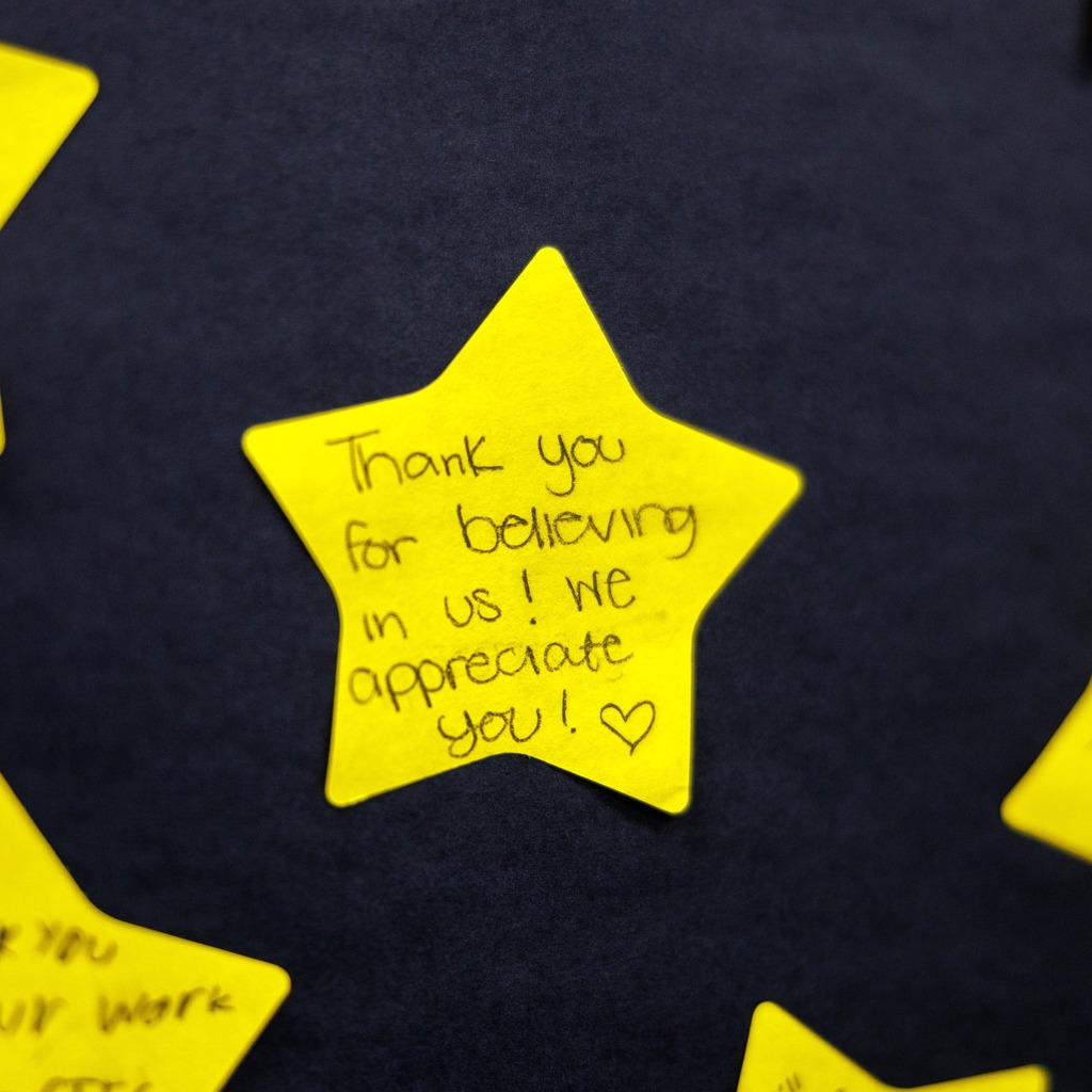 Close-up of a yellow, star-shaped note pinned to a dark bulletin board with a handwritten message that reads, ‘Thank you for believing in us! We appreciate you!’ followed by a heart. Other yellow star notes are partially visible around it.