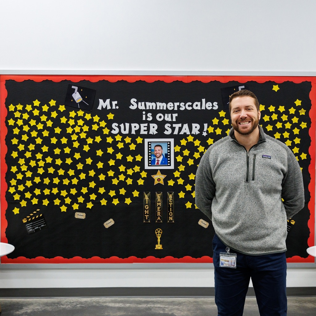 An adult staff member stands smiling in front of a school bulletin board decorated with hundreds of yellow star notes and the message ‘Mr. Summerscales is our SUPER STAR!’ The display includes movie-themed graphics, a photo of Mr. Summerscales, and award-style decorations recognizing him for outstanding contributions.