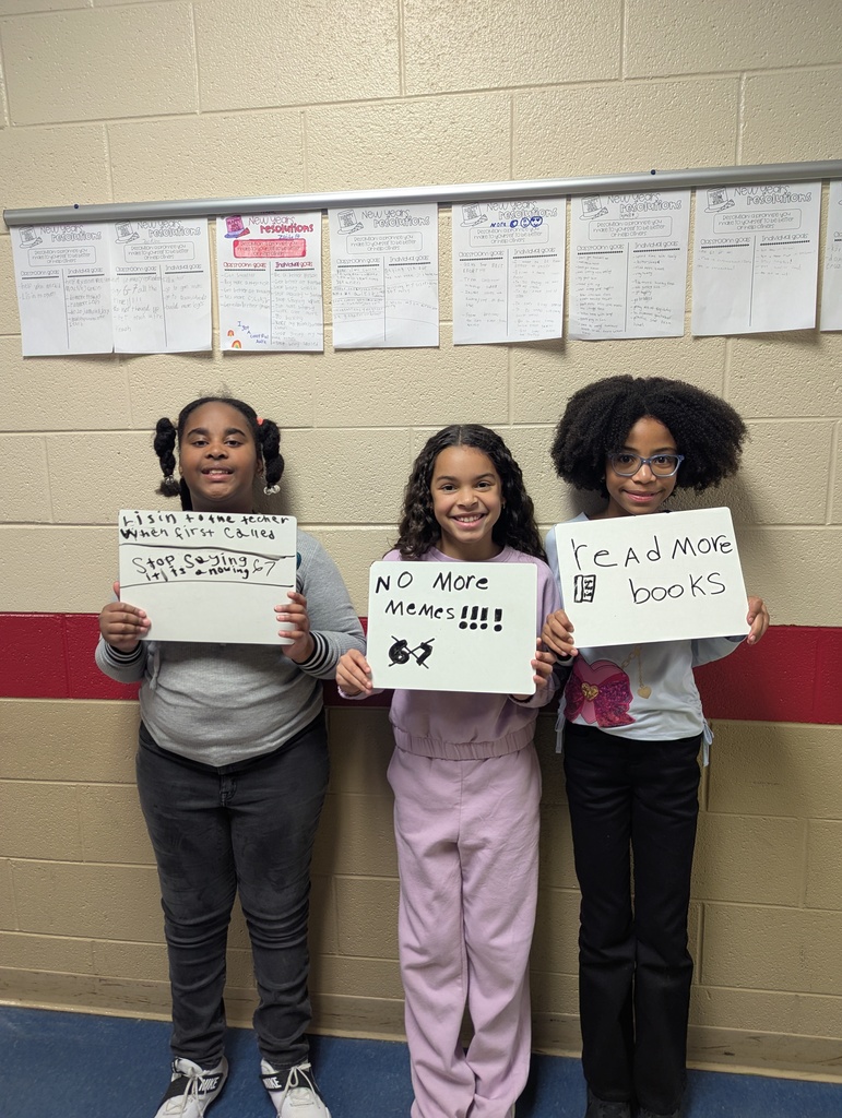 Three elementary students stand shoulder to shoulder holding whiteboards. The messages read “Listen to the teacher when first called,” “No more memes,” and “Read more books.” Student work labeled “New Year’s Resolutions” is displayed on the wall behind them.