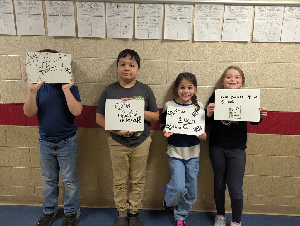 Four elementary students pose in a hallway holding whiteboards. The signs read “Say 6–7 all the time,” “Going to the shop in Reflex,” “Read 1,000 books,” and “Keep getting A+ in school.” The students smile proudly in front of a tan brick wall.