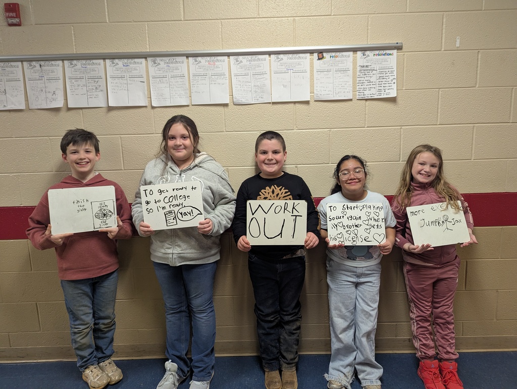 Five elementary students stand in a hallway holding whiteboards with goals such as “Finish the giver,” “To get ready to go to college,” “Work out,” “To start playing soccer again,” and “More exercise.” All students are smiling, with classroom writing posted above them.