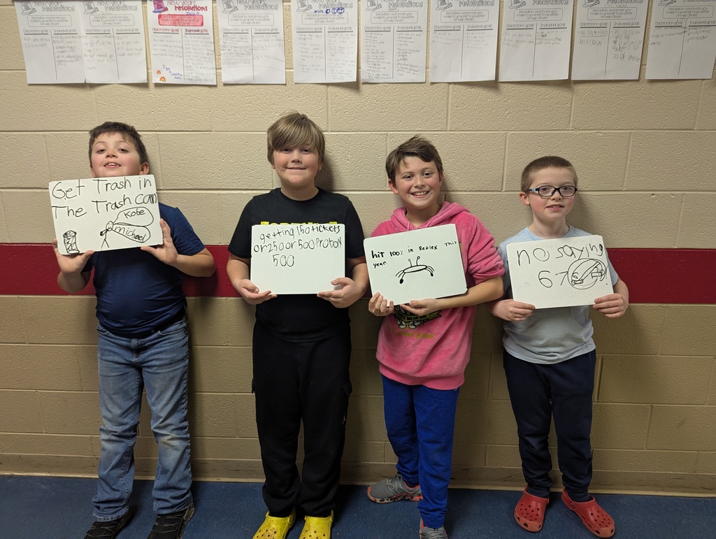 four elementary students line up in a hallway holding whiteboards with personal goals. Messages include “Get trash in the trash can,” “Getting 500 tickets,” “Hit 100% in Reflex this year,” and “No saying 6–7.” The students smile while standing in front of a tan brick wall.