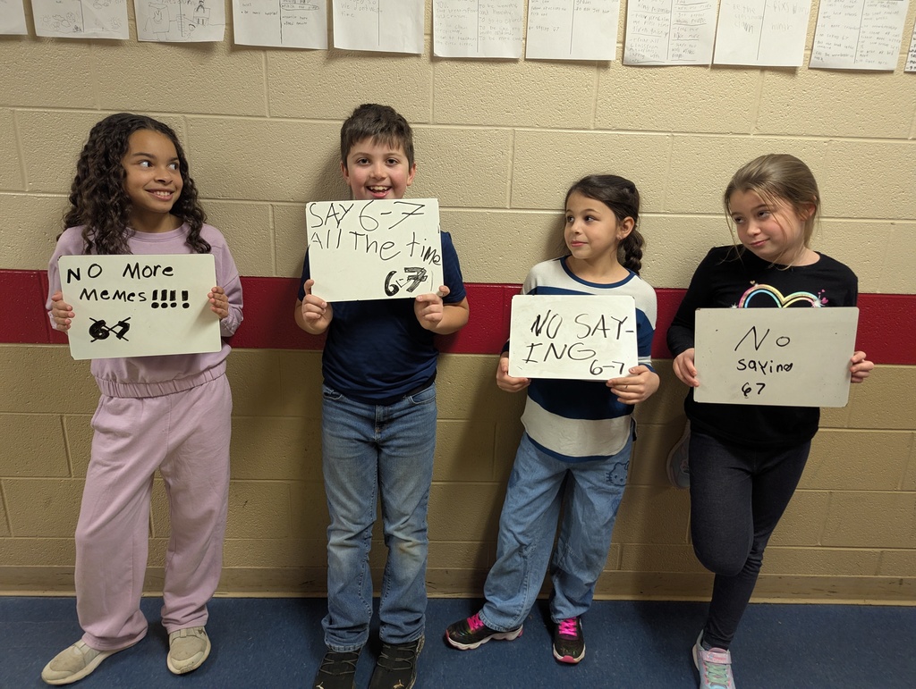 Four elementary students stand in a school hallway holding whiteboards with handwritten goals. The signs read “No more memes,” “Say 6–7 all the time,” and “No saying 6–7.” The students are smiling and standing against a tan brick wall with classroom papers displayed above them.