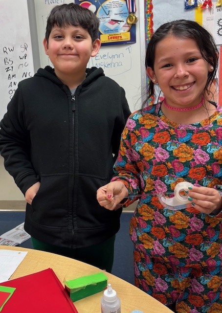 Two smiling students stand at a table holding small craft materials, including tape and cardboard pieces, as part of a classroom engineering design activity.