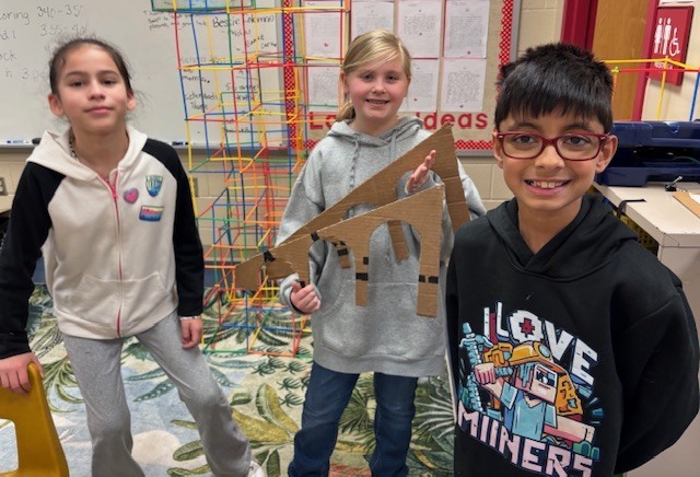 Three students stand and smile while holding a cardboard aqueduct structure with arches, showing their engineering design project in the classroom.
